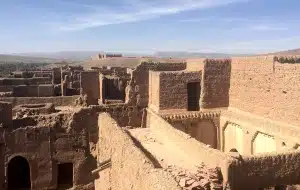 A traditional mud-brick Kasbah featuring intricate geometric patterns under a clear blue Moroccan sky, with greenery in the foreground.