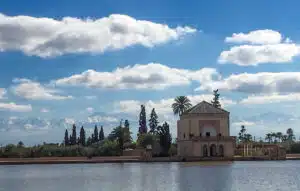 A pavilion with a green-tiled roof sits beside a large reflective body of water, surrounded by palm trees and mountains in the background, under a clear blue sky. This serene scene is to be found in the Menara Garden during our South of Morocco tour.