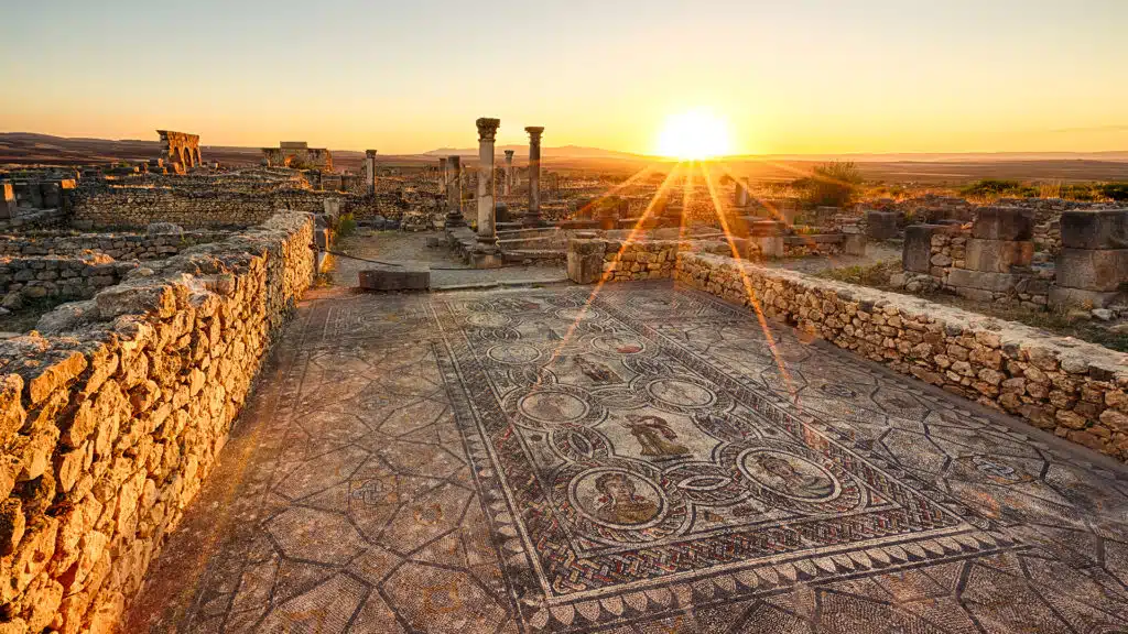 Sunset in the ancient ruins of Volubilis, one of Morocco's most prominent UNESCO historical sites, reminiscent of the grandeur of the Temple of Venus, with scattered stone remnants and greenery in the foreground.