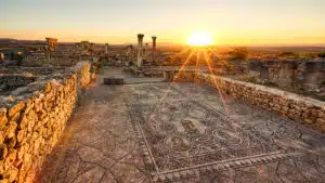 Sunset in the ancient ruins of Volubilis, one of Morocco's most prominent UNESCO historical sites, reminiscent of the grandeur of the Temple of Venus, with scattered stone remnants and greenery in the foreground.