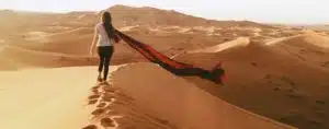 Traveler walking along a sand dune in the Moroccan desert, with a flowing scarf trailing behind, experiencing the serene beauty of the Sahara on a private tour of Morocco.
