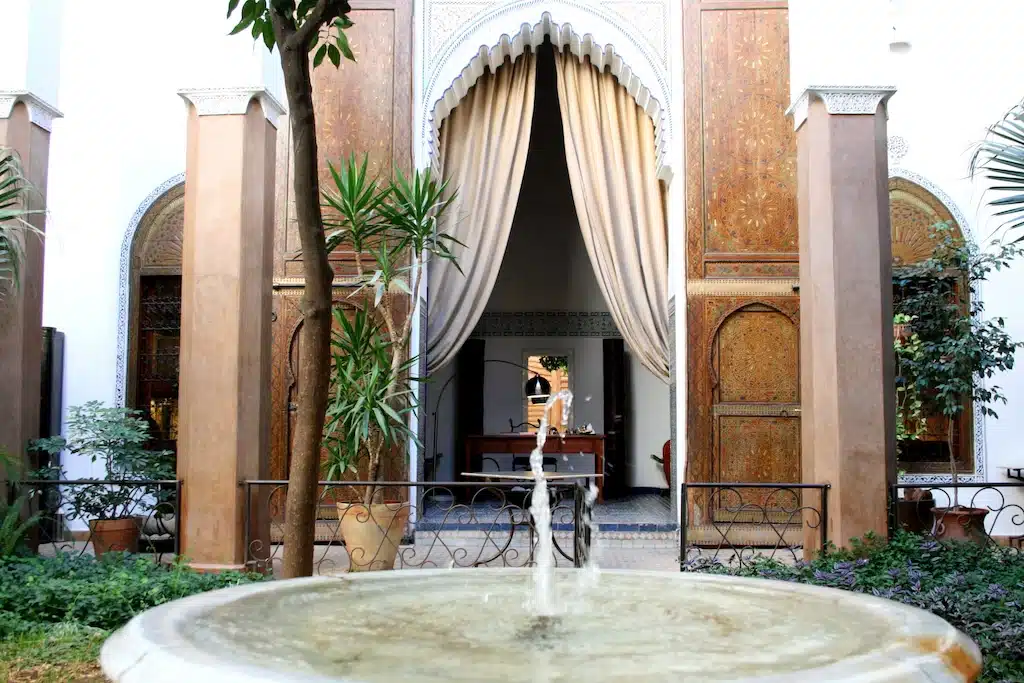 a typical riad in Morocco displaying a garden with a fountain in the foreground and a view of a cosy room in the background