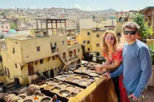 a couple of tourists smiling into the camera while watching a Moroccan Medina from a balcony