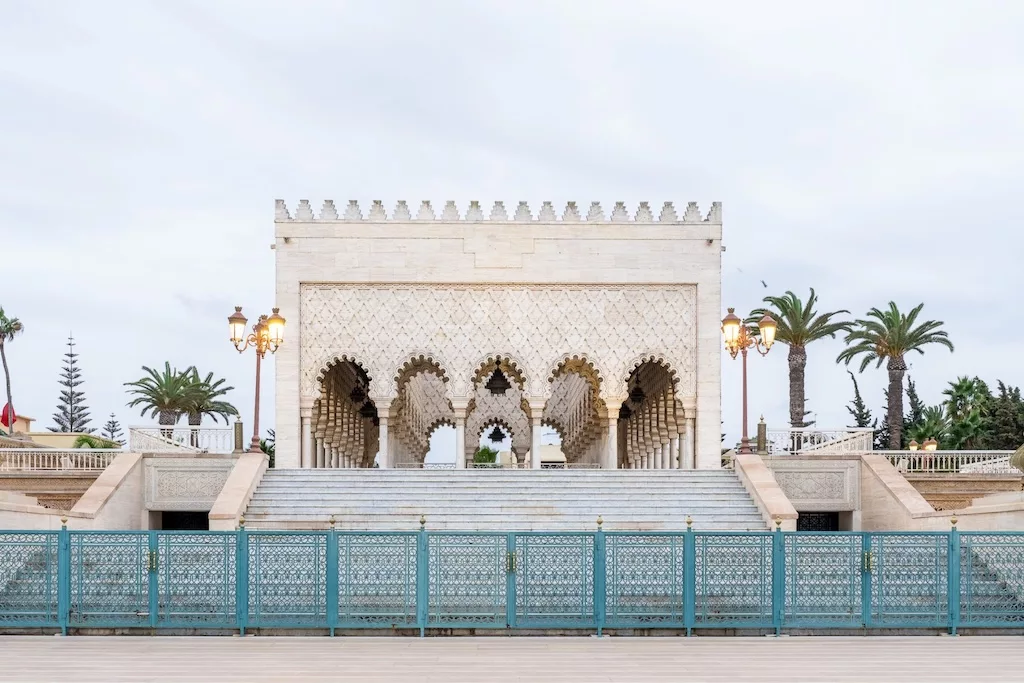 A marble mausoleum with arched entryways and intricate designs, flanked by palm trees and ornate lamp posts, sits atop a series of steps behind a decorative teal fence. This architectural gem is one of the highlights you might encounter on Morocco tours through Rabat, showcasing the city's rich heritage.