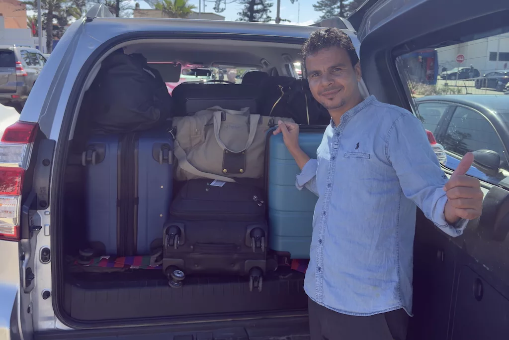 a male traveler standing in front of an open car trunk filled with suitcases