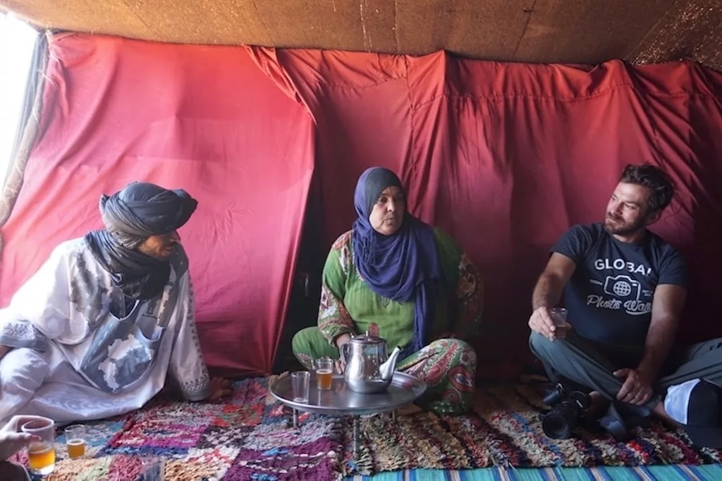 a tourist sharing a cup of tea with a local Moroccan woman and his local guide inside a beduin tent