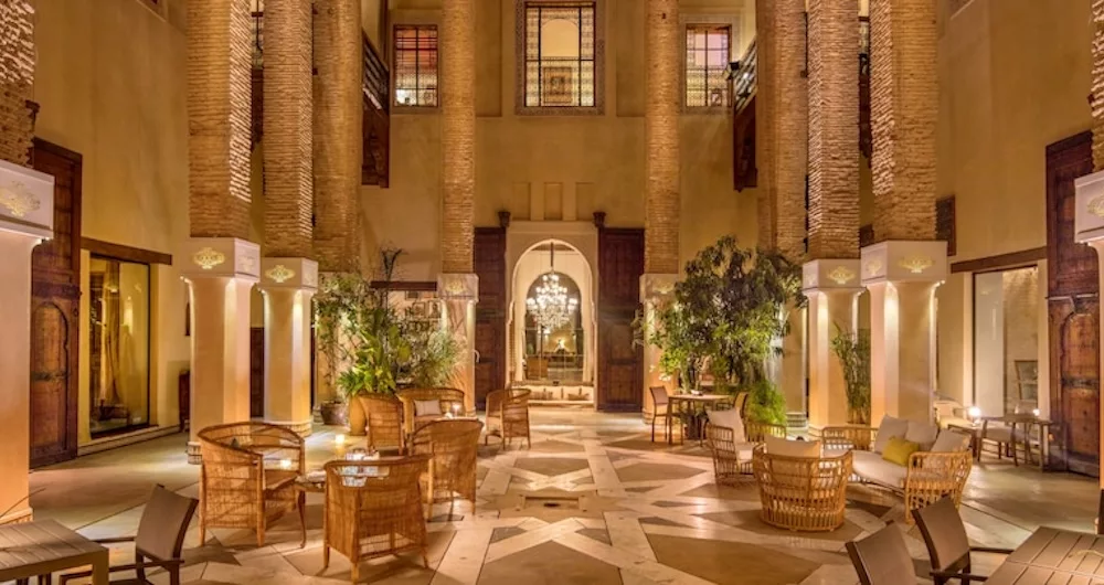 a well-lit and luxurious-looking courtyard with wooden chairs and tables in a high-class Moroccan riad