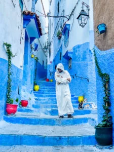 a Moroccan man in white clothing walking down the stairs in the blue city of Chefchaouen holding a cat