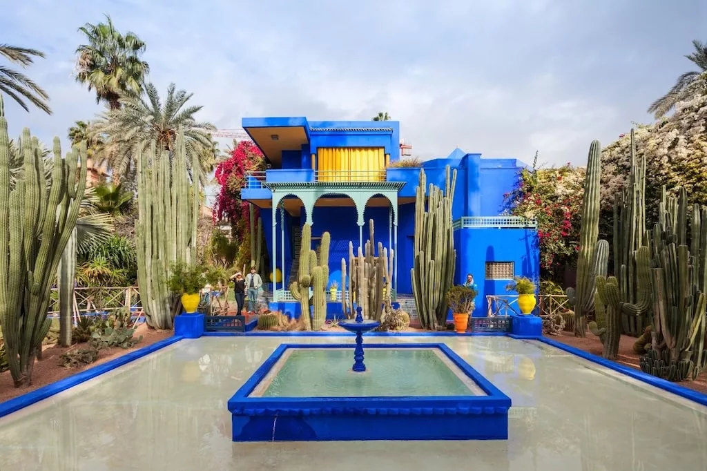 inside the Majorelle Garden in Marrakech, with cacti growing in abundance around a square fountain with a blue building behind it