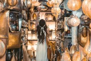 A female tourist in a white dress standing inside a small shop in the souks with silver goods, pointing at one of the silver lamps on the ceiling.