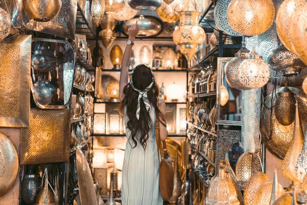 A female tourist in a white dress standing inside a small shop in the souks with silver goods, pointing at one of the silver lamps on the ceiling.