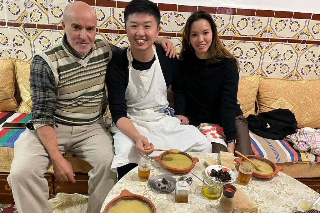 A young couple of travelers sharing lunch with Moustafa, a local Moroccan man that loves welcoming people from other cultures in his home.