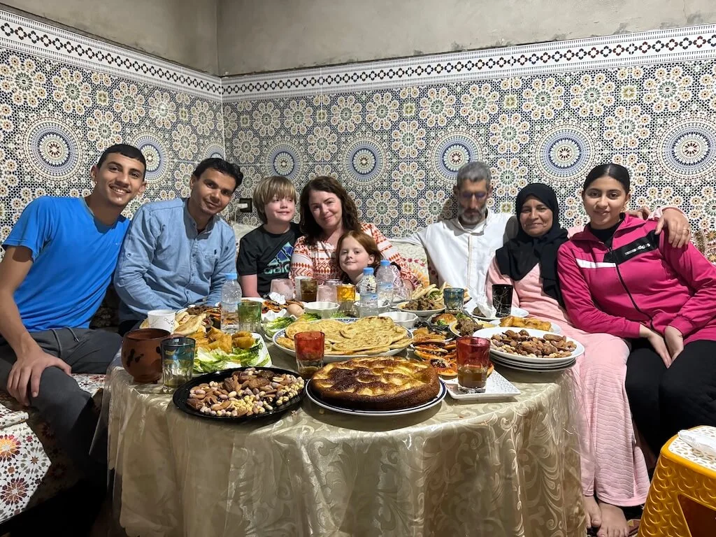 A family of tourists sharing iftar with a local family in Marrakech during Ramadan, posing together for a picture behind a able full of traditional home-cooked Moroccan food.