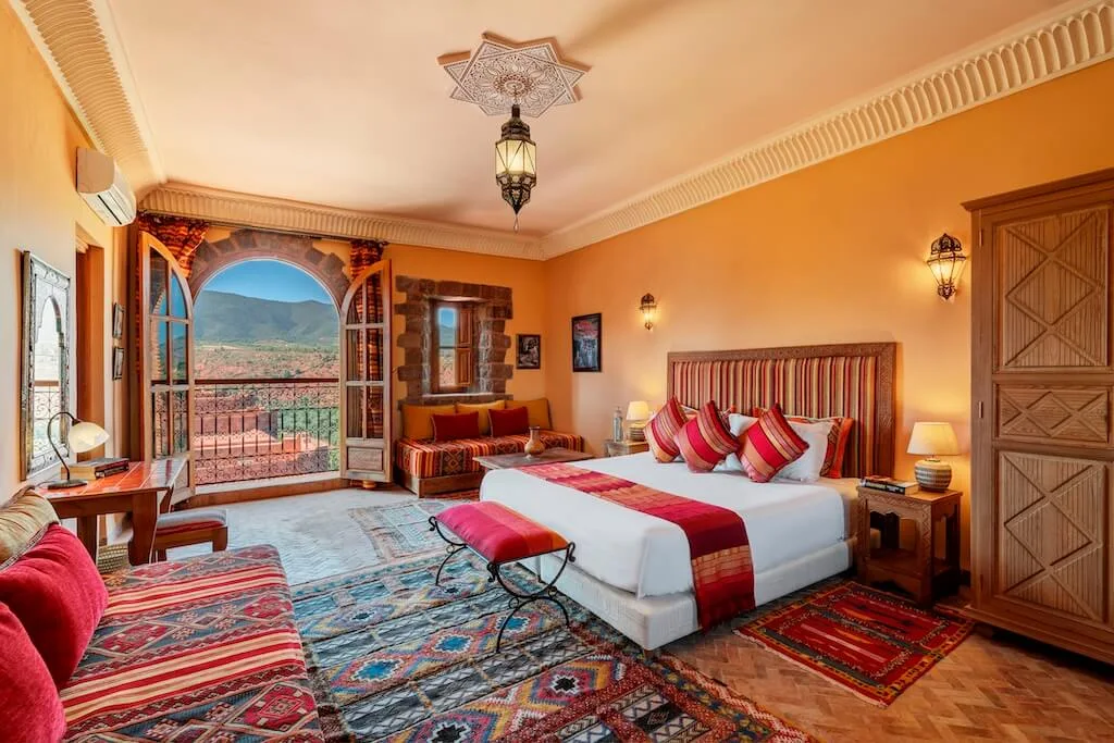 Inside a cozy room in Kasbah Angour with colorful rugs and an open door leading to a balcony with views of the kasbah.