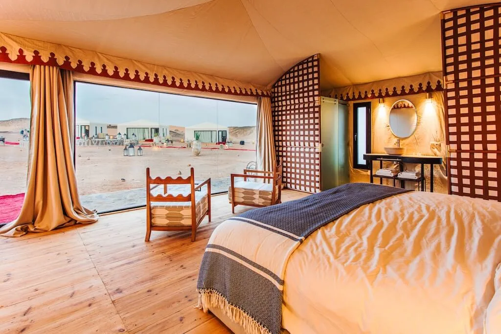 Inside a luxury tent in the dessert, with king-size bed, private bathroom and a seating facing a huge window with views of the desert.