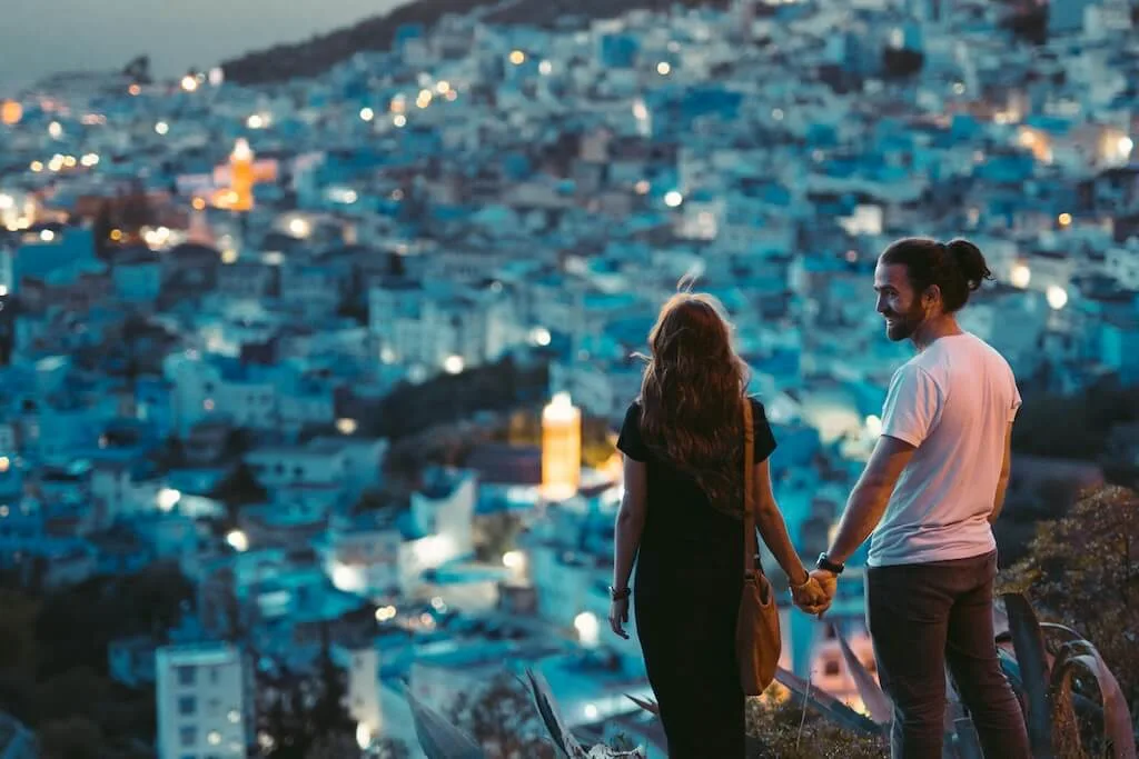 A couple holding hands while looking down from a viewpoint at the blue city of Chefchaouen in the evening light.