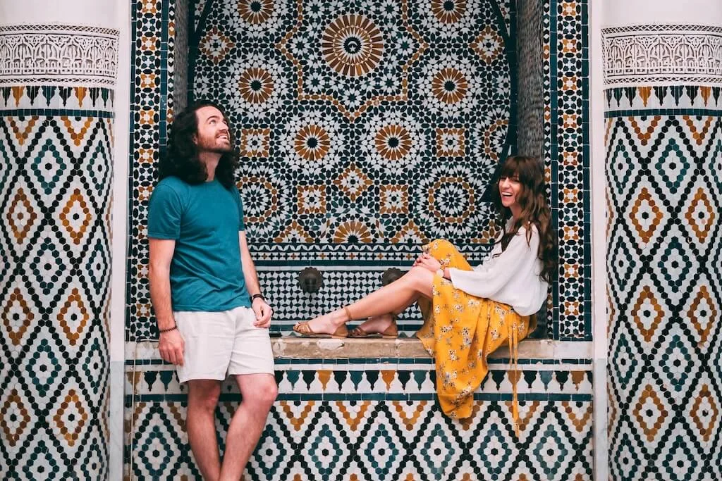 A young couple posing for a picture in front of an intricately decorated wall with traditional Moroccan tiles.