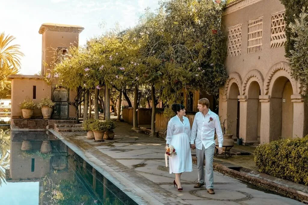 A couple on their honeymoon in Morocco strolling around a swimming pool inside a historic kasbah.