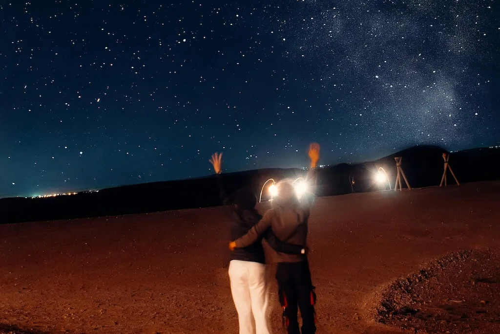 A couple hugging in the Sahara desert under a starry night sky.