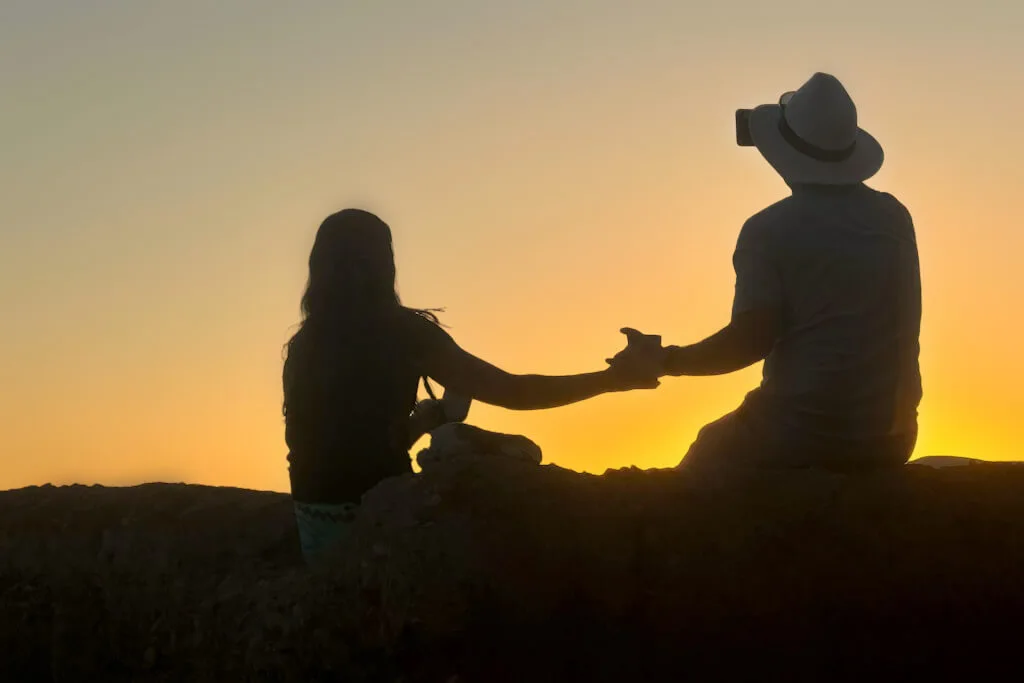 A couple sharing a glass of wine on the dunes in the Sahara desert with the sun setting in the background.