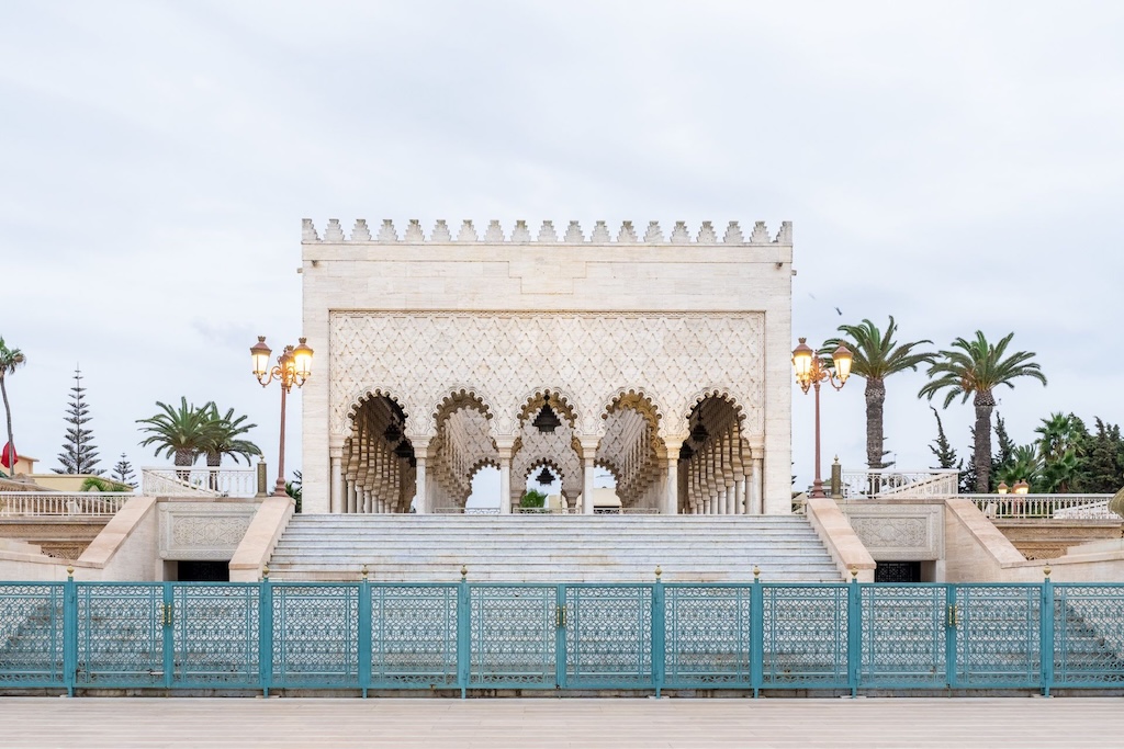 A marble mausoleum with arched entryways and intricate designs, flanked by palm trees and ornate lamp posts, sits atop a series of steps behind a decorative teal fence. This architectural gem is one of the highlights you might encounter on Morocco tours through Rabat, showcasing the city's rich heritage.