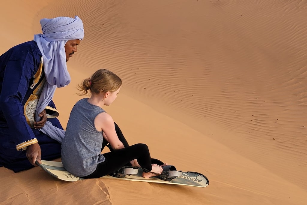 A Moroccan man pushing a little girl seated of a sandboard down a dune in the Moroccan desert.