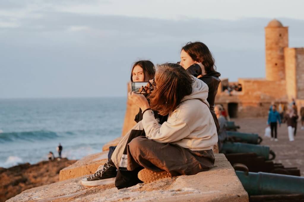 A group of female tourists sitting on the coastal walls of the Essaouira fortress taking a picture of each other.